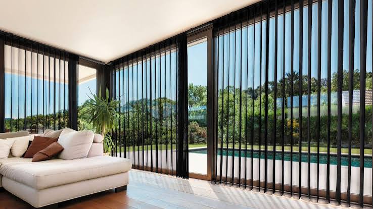 Sheer vertical SmartDrape fabric vanes by Norman gliding across floor-to-ceiling sliding glass doors overlooking a pool in a luxury Sarasota living room by Sarasota Window Blinds